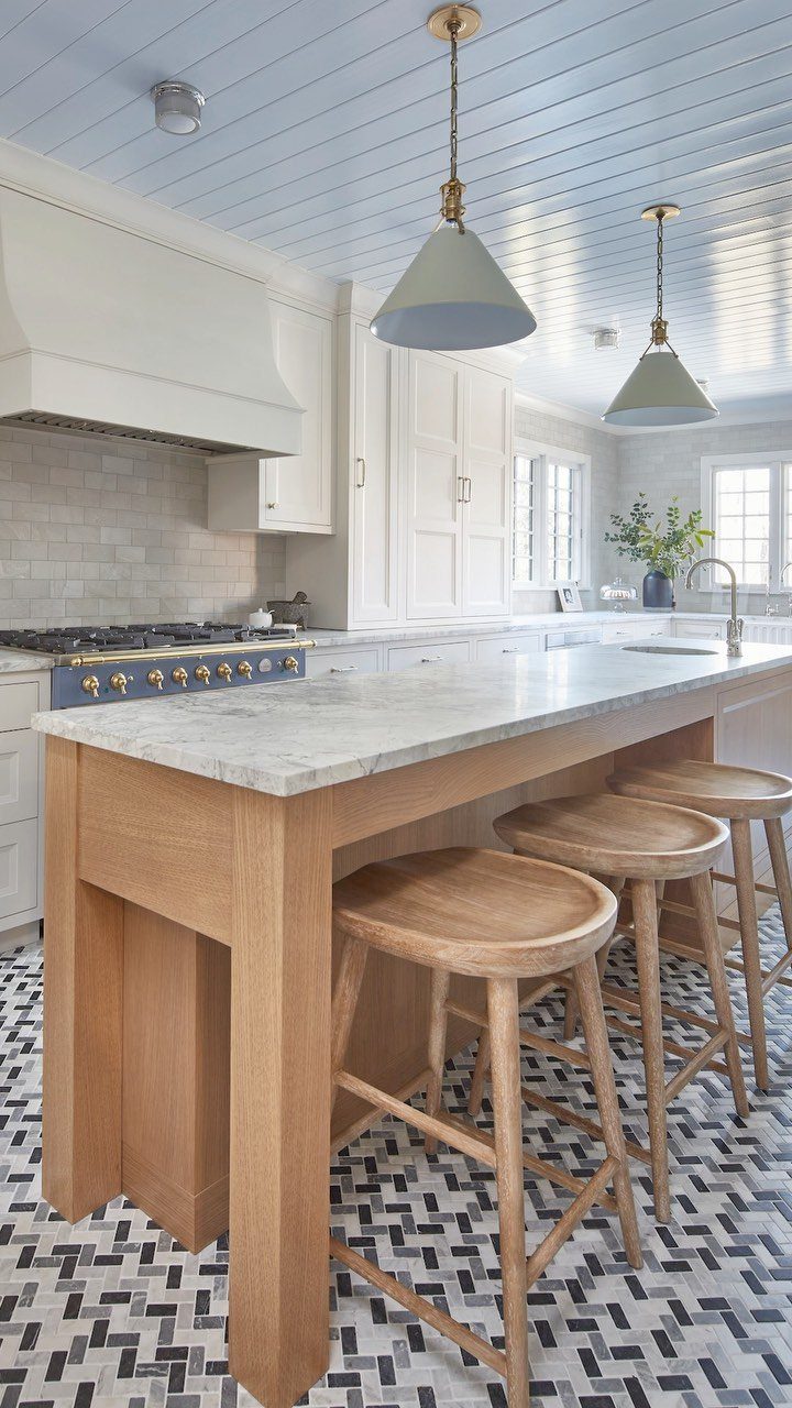 A narrow wooden kitchen island with a marble top and three wooden stools in a bright kitchen.