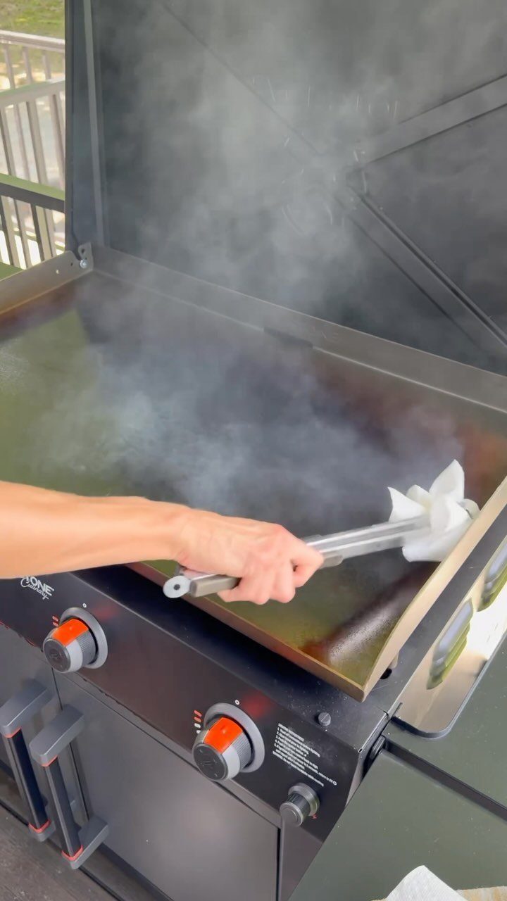 A hand uses a cloth tool to wipe oil onto a steaming hot griddle surface.