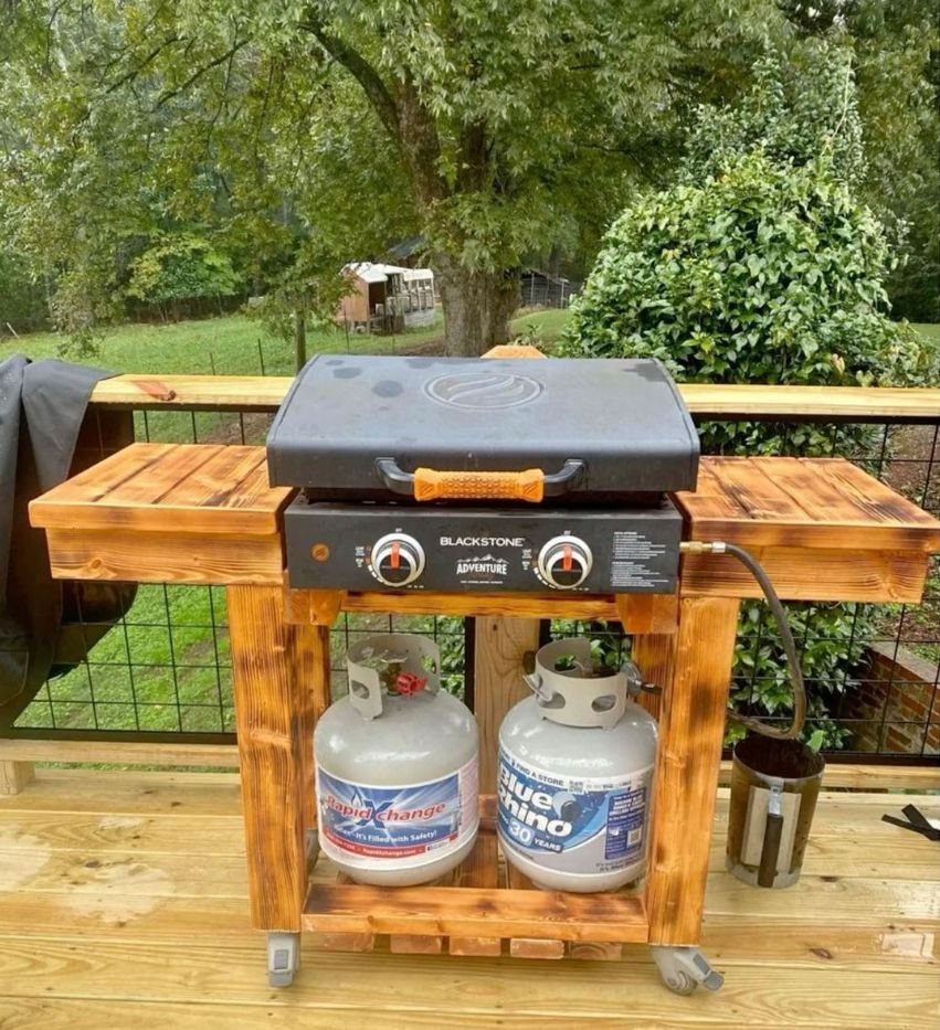 A wooden cart holding a Blackstone griddle with two propane tanks underneath on a deck.