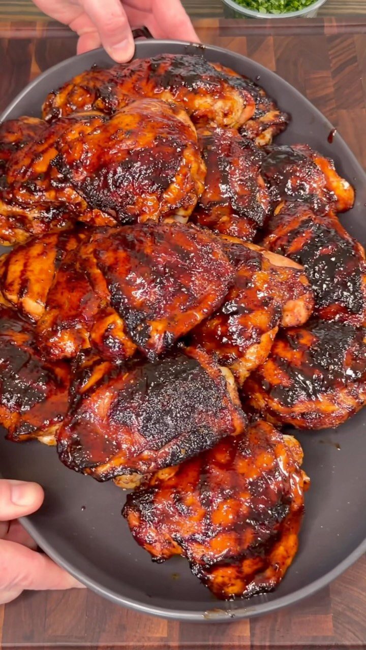 Glazed grilled chicken thighs piled on a dark grey platter held by hands.