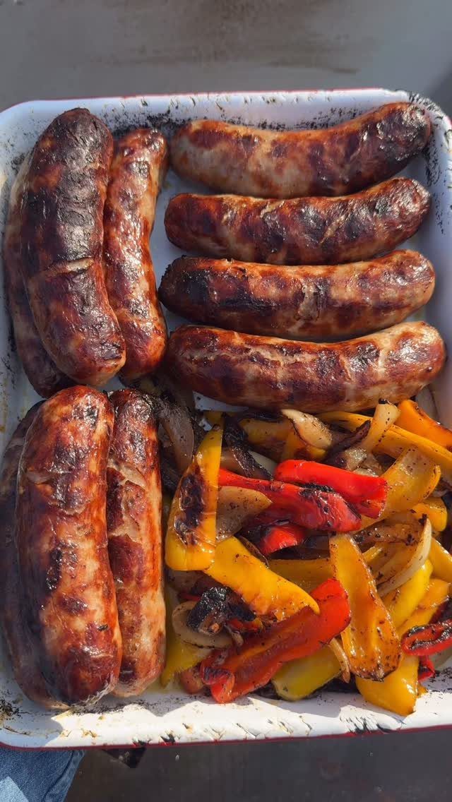 Grilled sausages and sliced bell peppers with onions in a white baking dish.