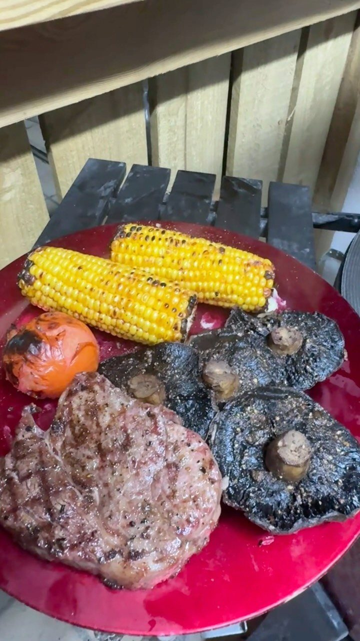 Grilled steak served on a red plate with corn, tomato, and large mushrooms.