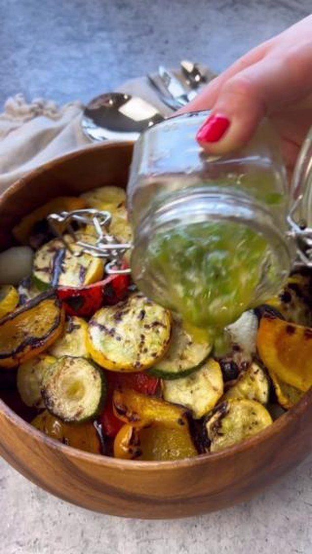 Hand pouring green dressing over a wooden bowl of grilled zucchini and summer squash.