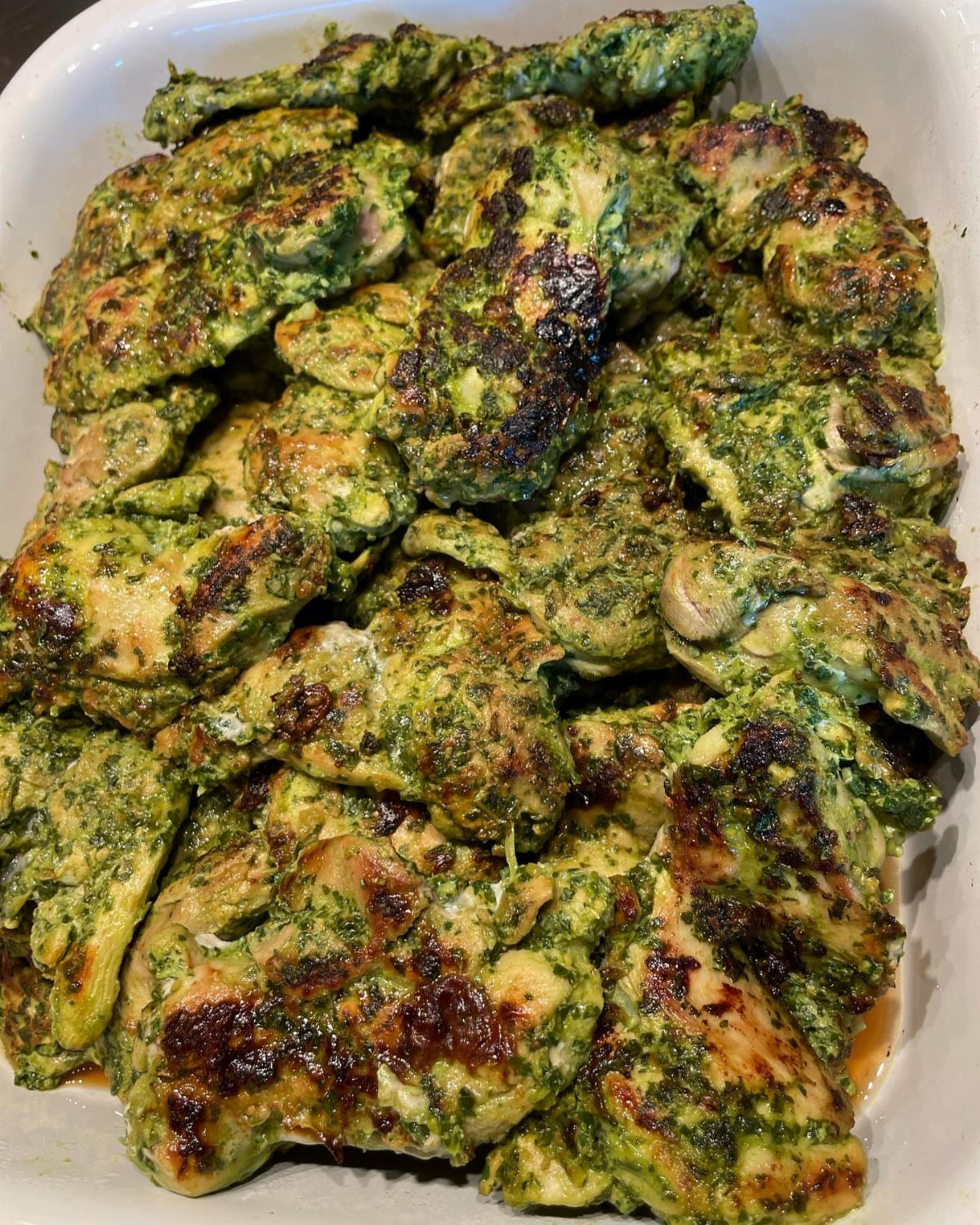 Grilled chicken pieces coated in green pesto sauce served in a white dish.