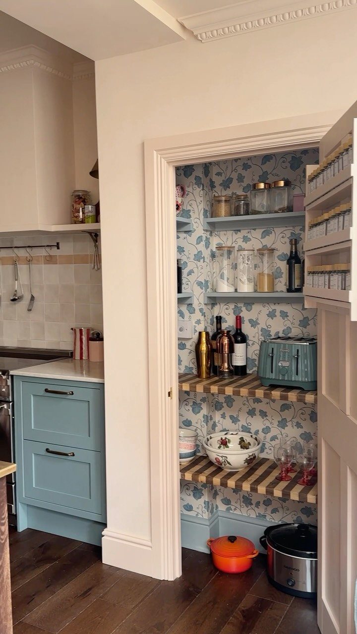 A small larder with blue floral wallpaper, open shelving, and wooden butcher block shelves containing kitchen supplies.