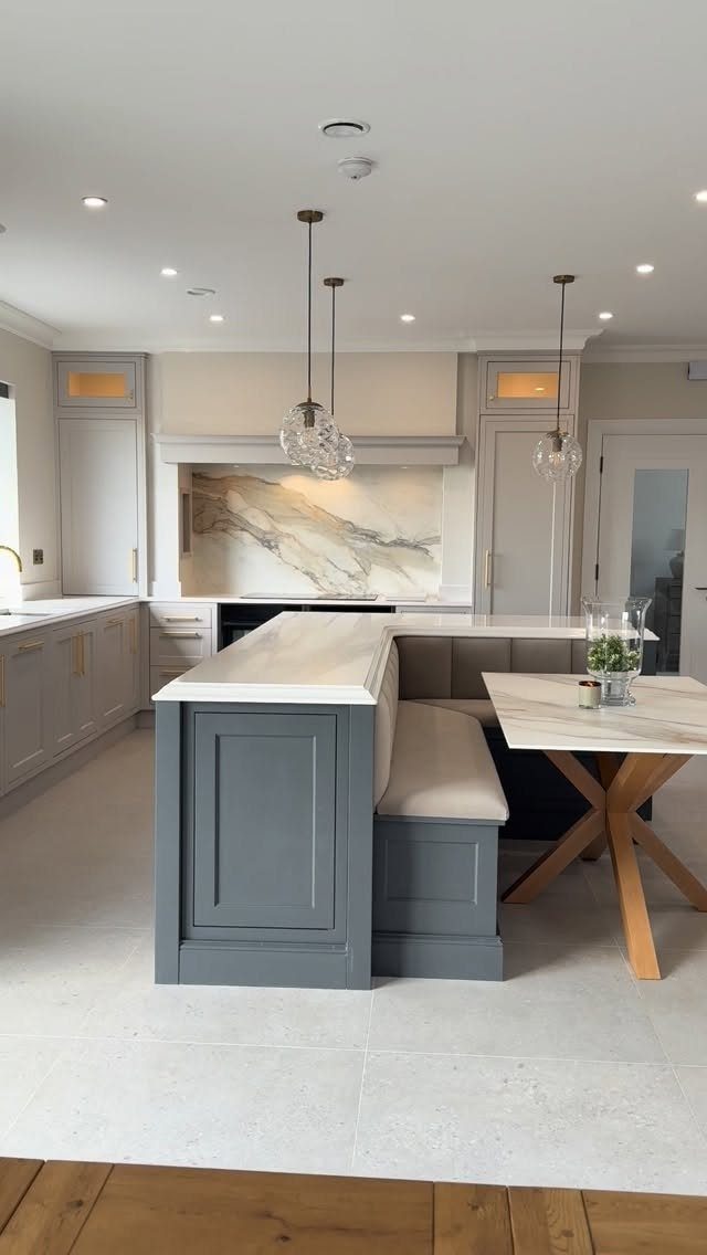 Grey kitchen island with white marble countertop and built-in banquette seating under glass pendant lights.