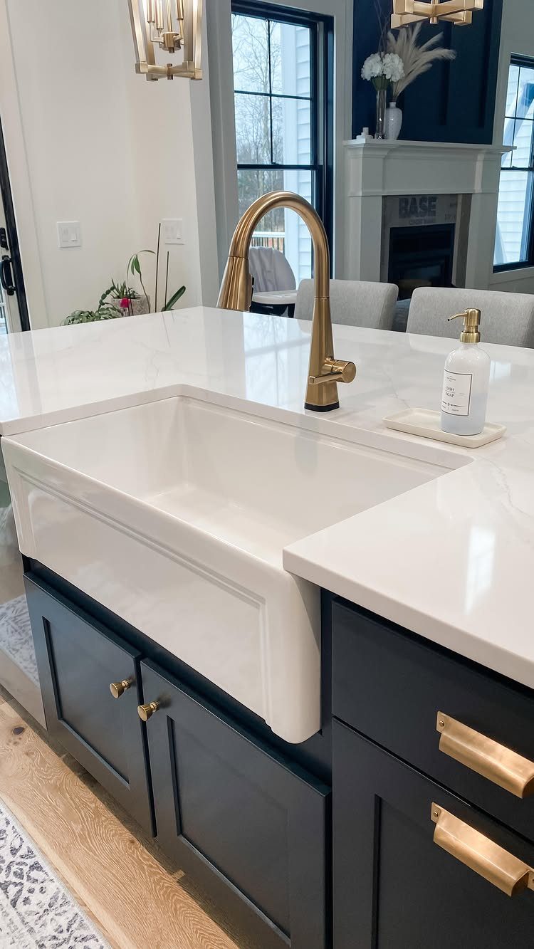 White apron-front farmhouse sink installed in a dark blue kitchen island with gold hardware.