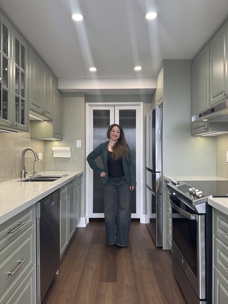 Green Kitchen Cabinets: 12 Ideas for Every Shade 3 A woman standing in a kitchen with sage green cabinets and white countertops.