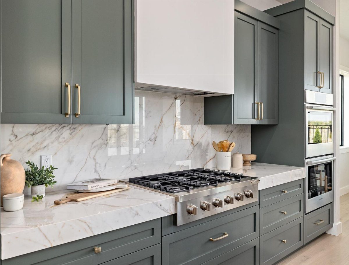 Green Kitchen Cabinets: 12 Ideas for Every Shade 9 Dark green kitchen cabinets with gold handles and marble countertops surrounding a stainless steel stove.