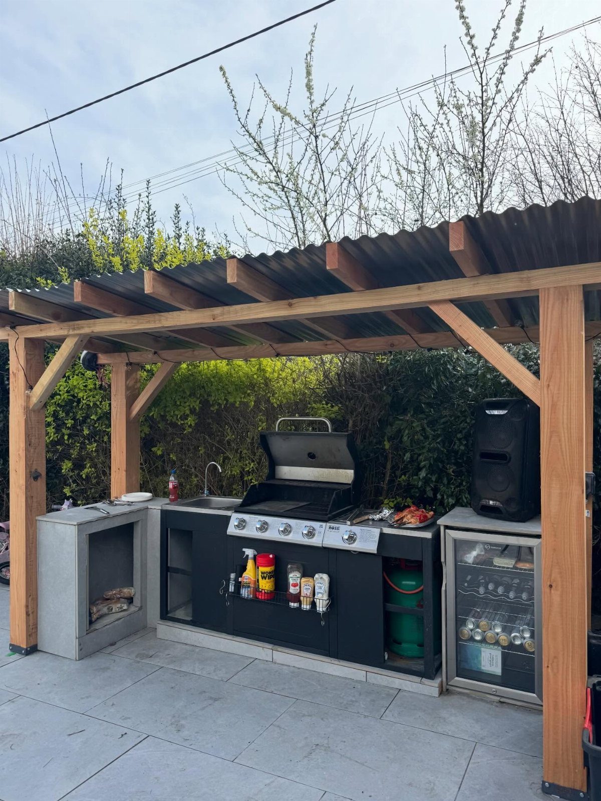 A wooden pergola with a corrugated roof shelters a black outdoor kitchen with a built-in grill.