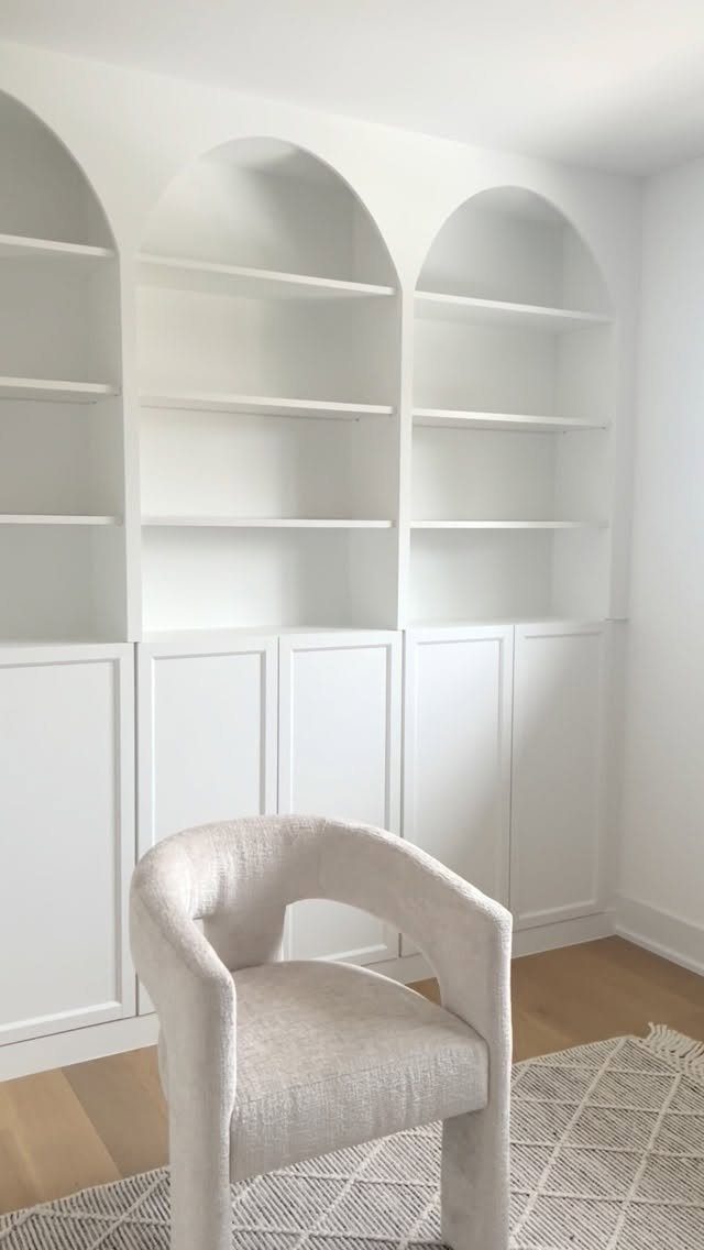 White built-in arched shelving units with bottom cabinets and a white chair in a bright room.
