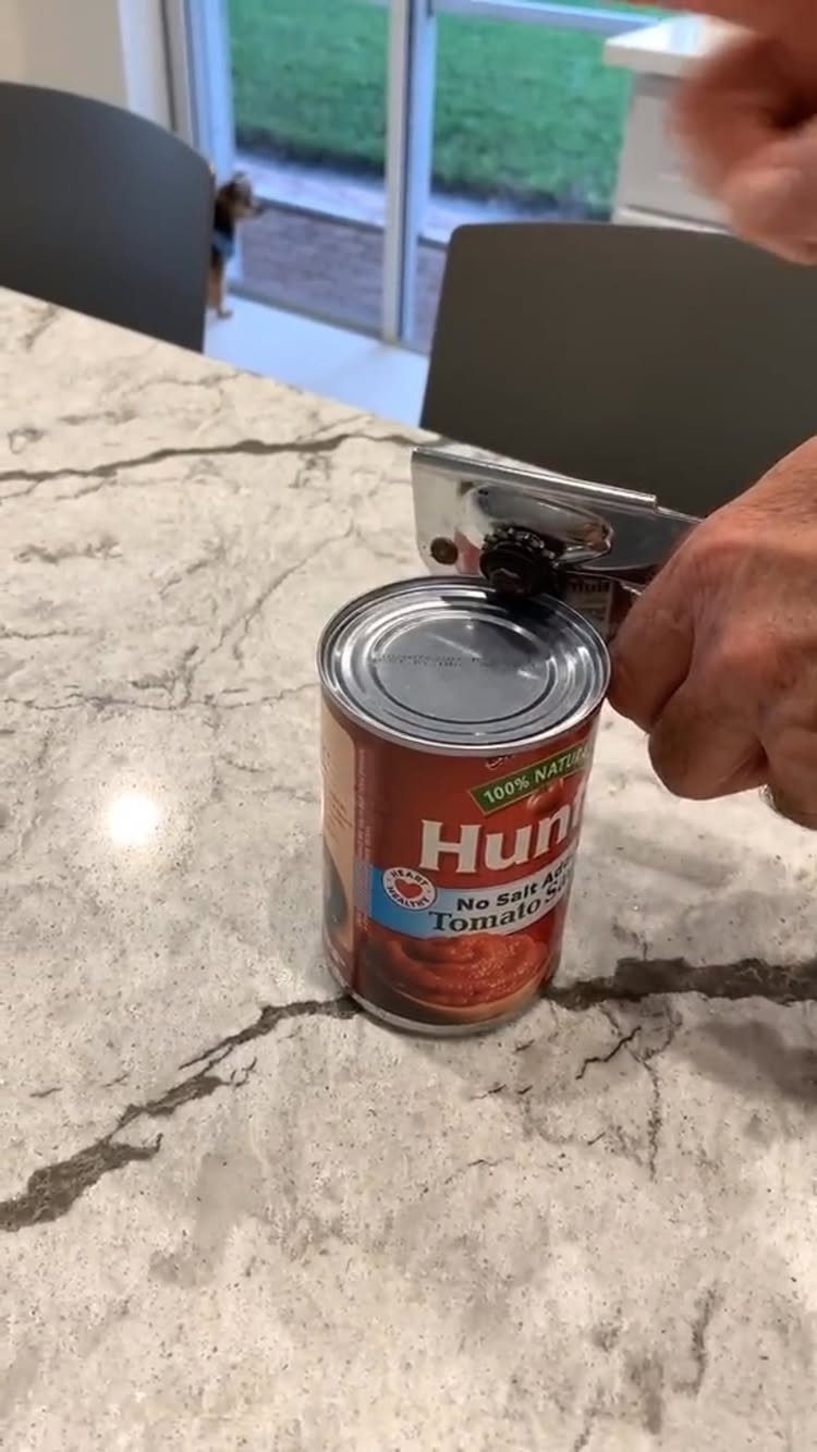 Hands using a metal can opener to open a red can of tomato sauce on marble.