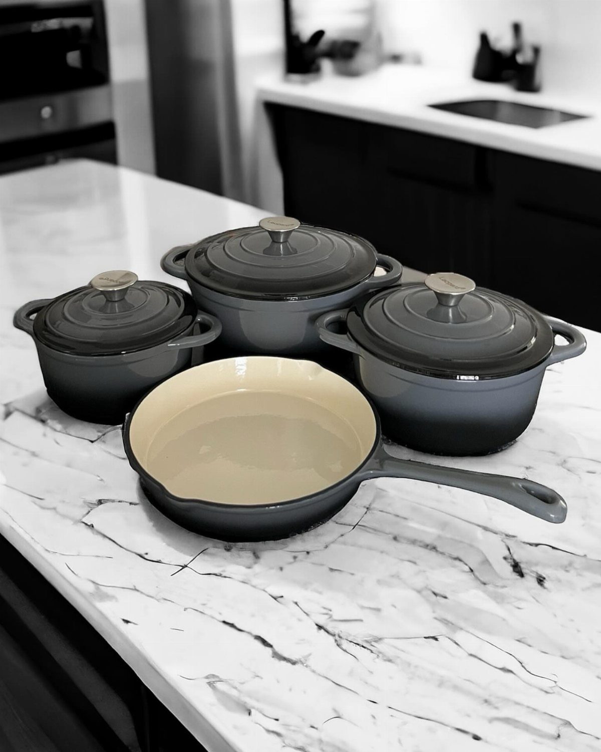 Enameled cast-iron cookware including three lidded pots and one large skillet on a marble countertop.