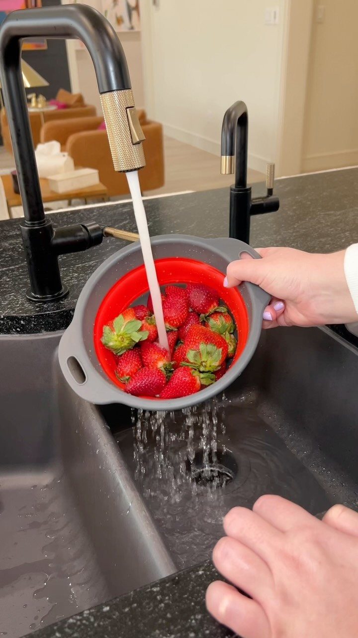 A person rinsing bright red strawberries in a grey and red colander under a running kitchen faucet.