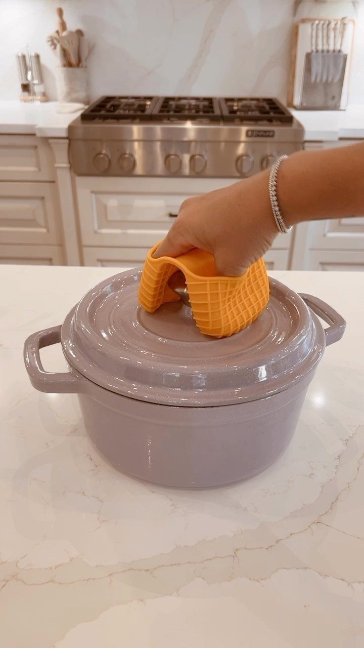 A hand using an orange silicone potholder to lift the lid of a purple Dutch oven.