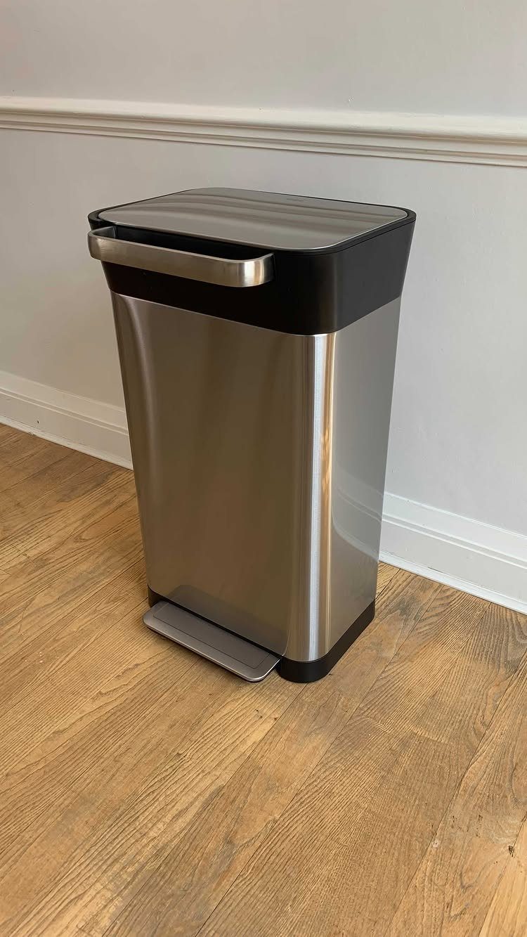 A stainless steel step trash can with a black lid on a wooden floor.
