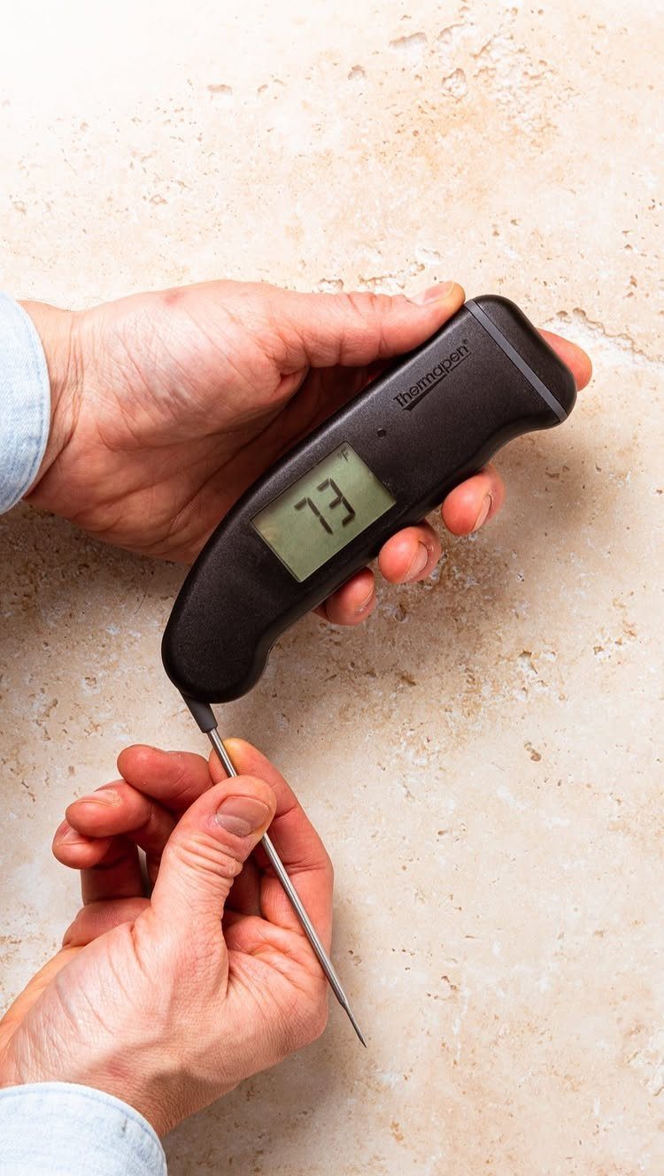 Hands holding a black digital instant-read thermometer with a metal probe over a stone surface.