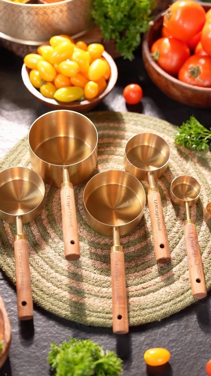 Gold metal measuring cups with wooden handles resting on a woven mat surrounded by fresh vegetables.