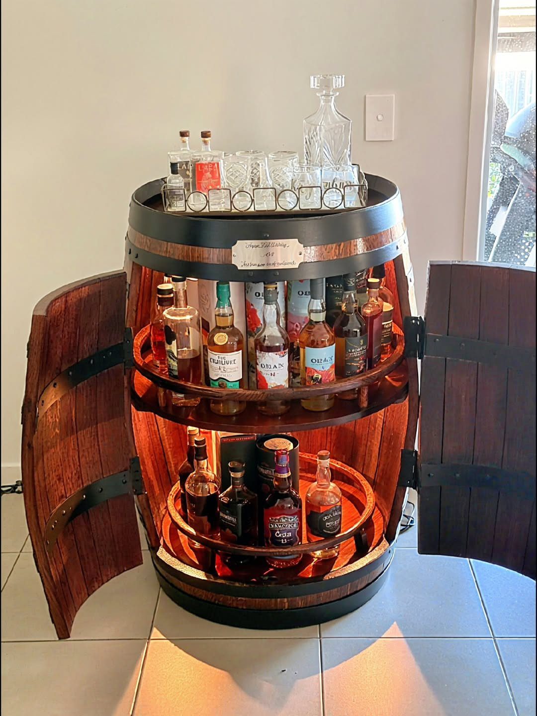 A wooden barrel bar cabinet with open doors displaying various liquor bottles and glassware.