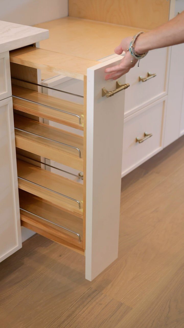 A hand pulls out a narrow wooden pull-out organizer from a white kitchen cabinet.
