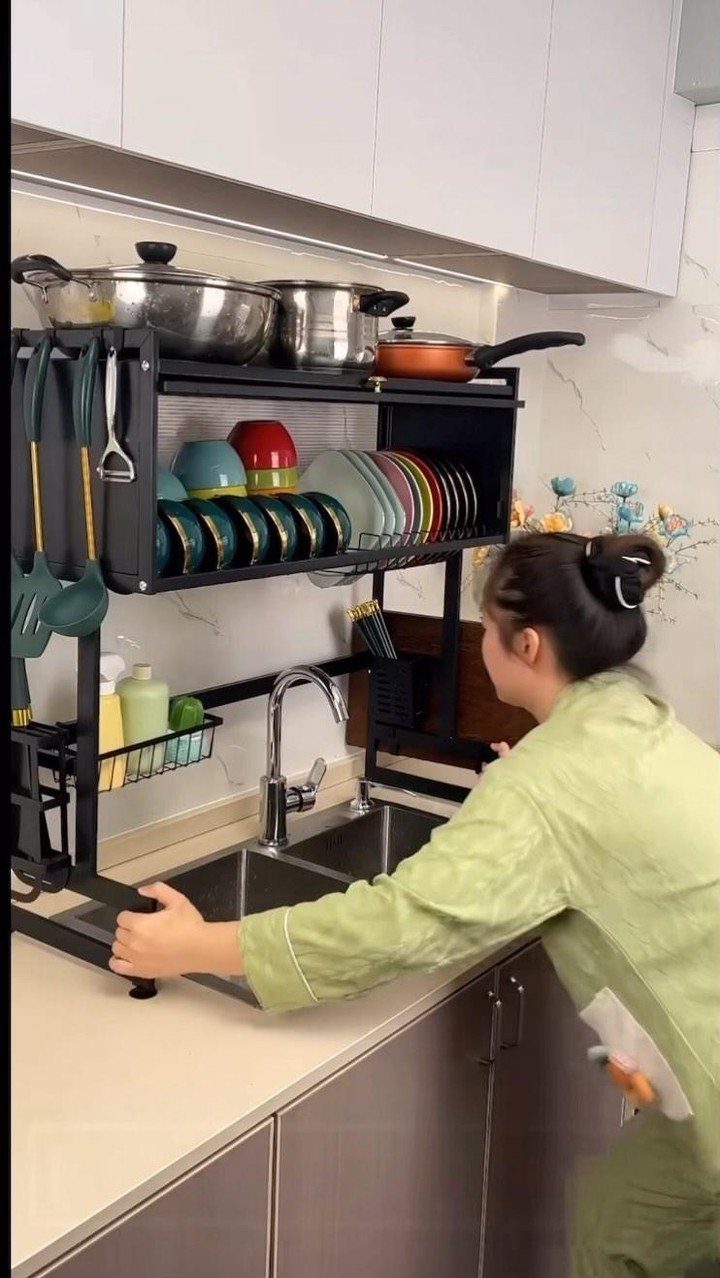 A person placing a black multi-tier metal organizer rack over a kitchen sink.