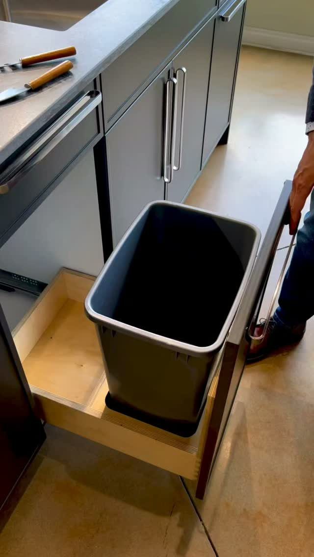 A black trash bin inside an open wooden pull-out drawer in a gray kitchen cabinet.