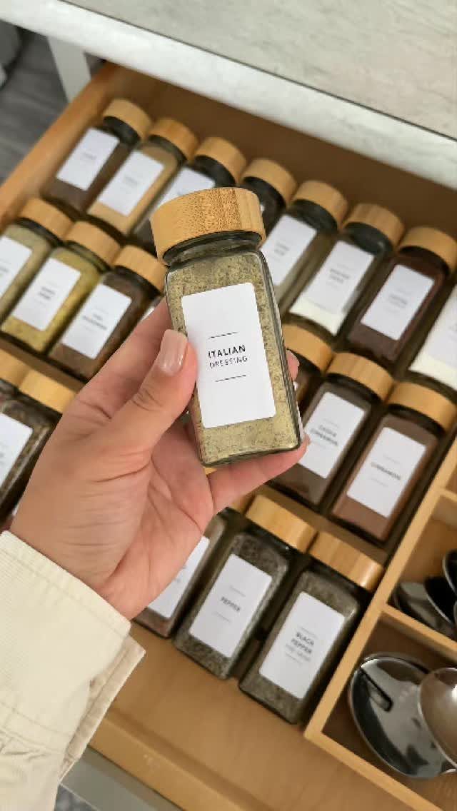 A hand holding a glass spice jar labeled Italian Dressing over an organized wooden drawer.