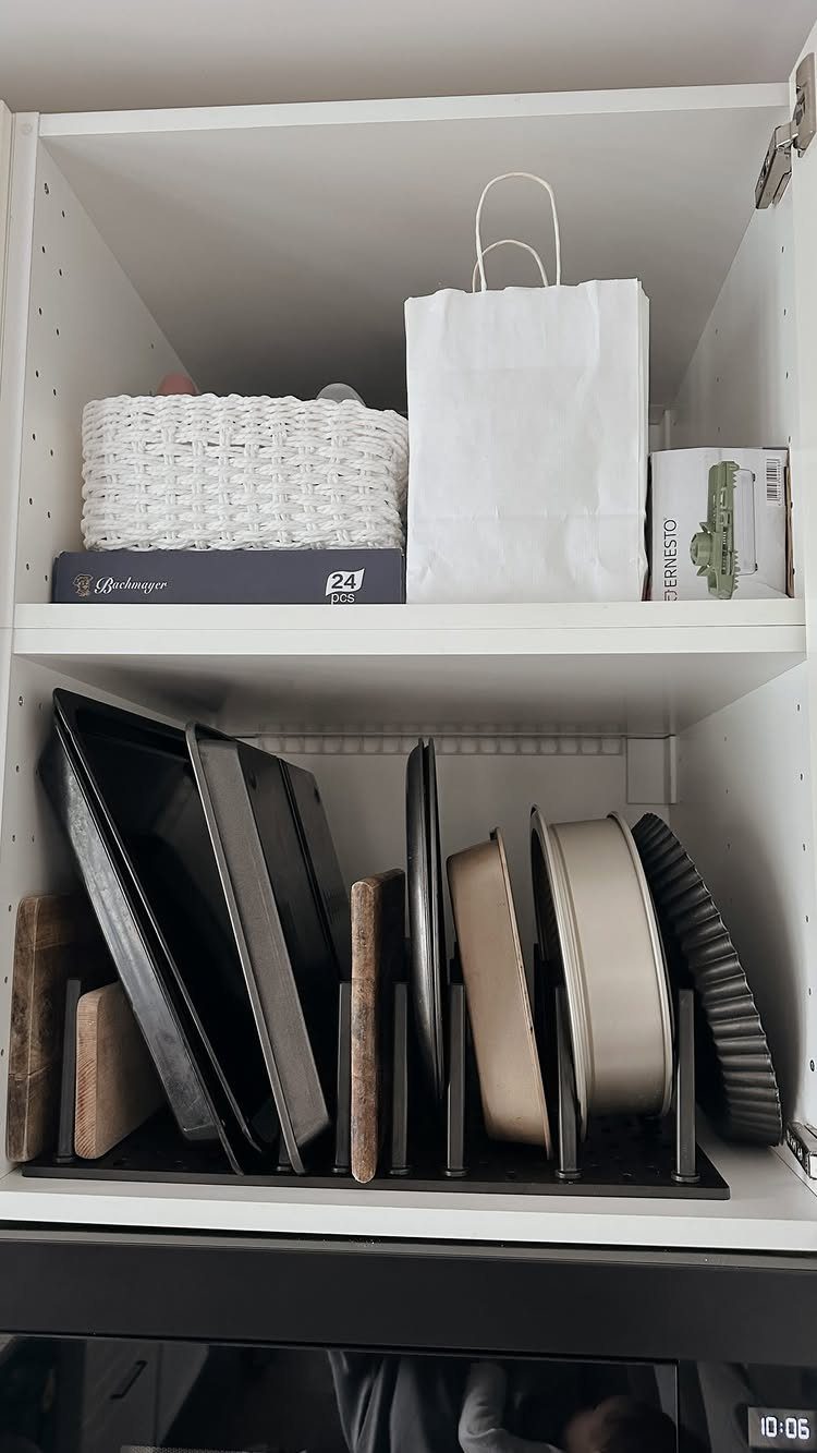 Baking sheets and pans stored vertically in a black organizer inside a white kitchen cabinet.