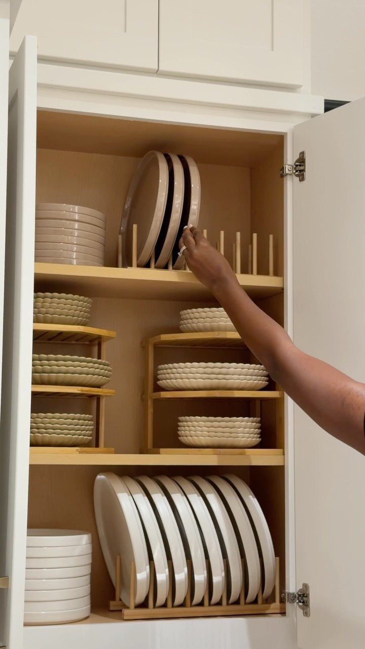 A person reaching into a white kitchen cabinet to organize white plates using wooden shelf organizers.