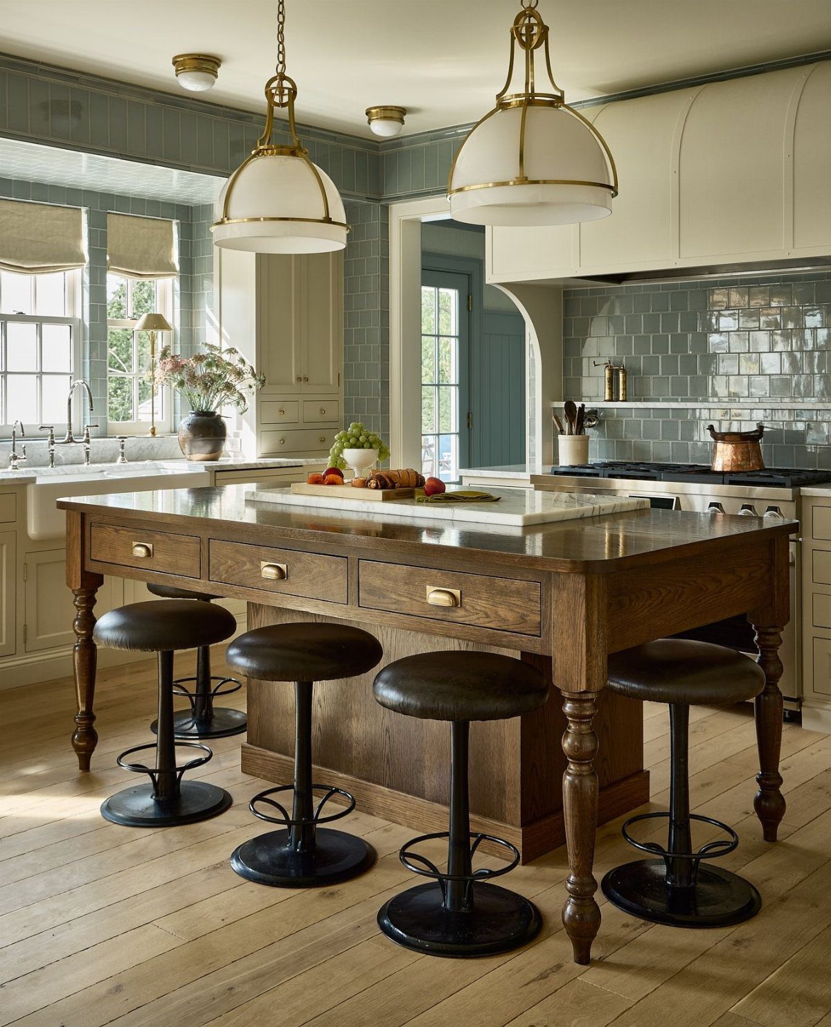 A wooden baker's table with drawers and four leather stools in a bright kitchen.