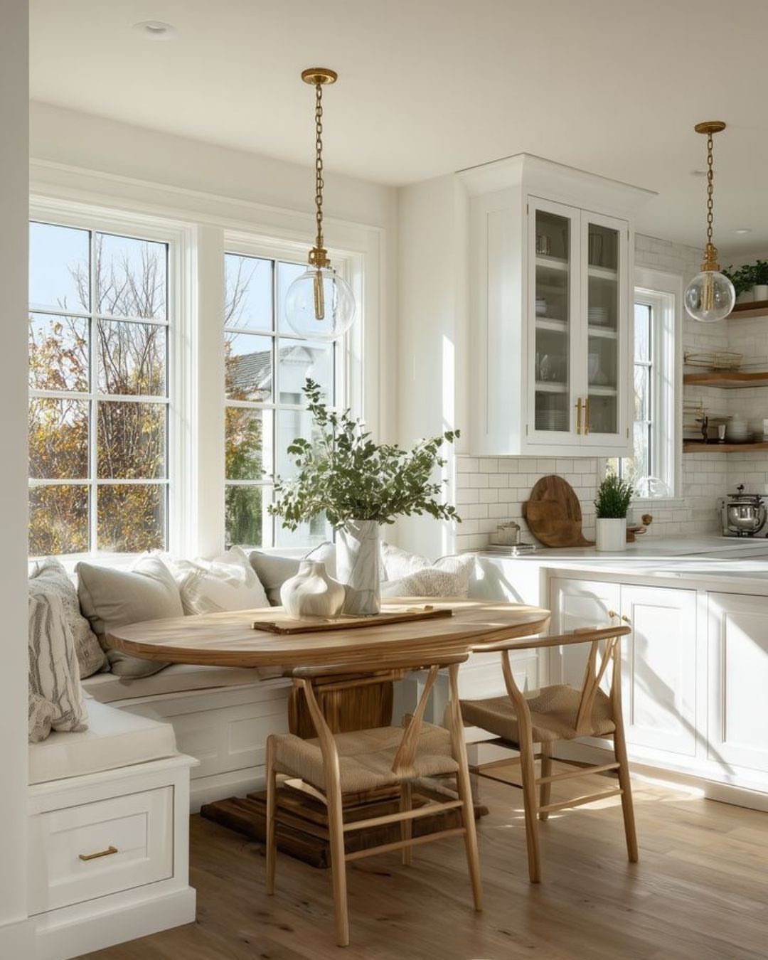 A wooden dining table and built-in banquette seating situated by large bright windows.