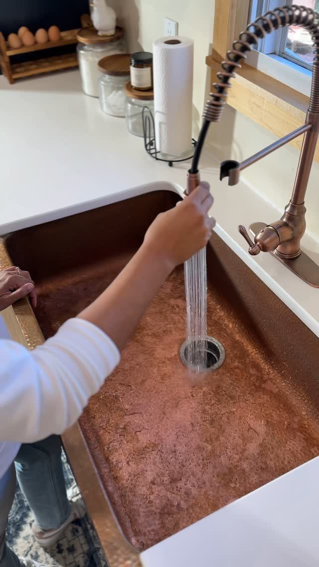 A person rinsing a large copper farmhouse sink with a high-arc kitchen faucet.