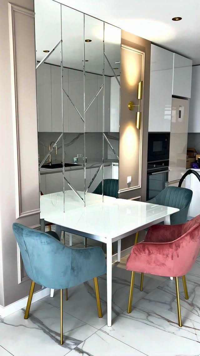 Geometric mirrored panels on a wall behind a white dining table and colorful velvet chairs.