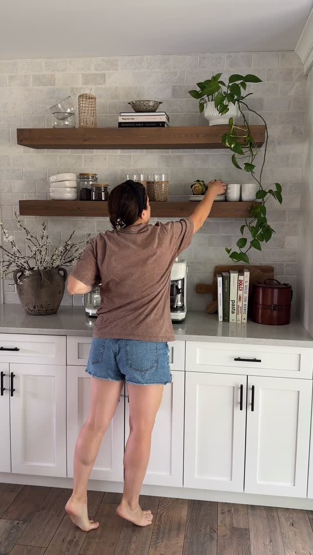 A woman reaching for a white bowl on wooden open shelves in a bright kitchen.