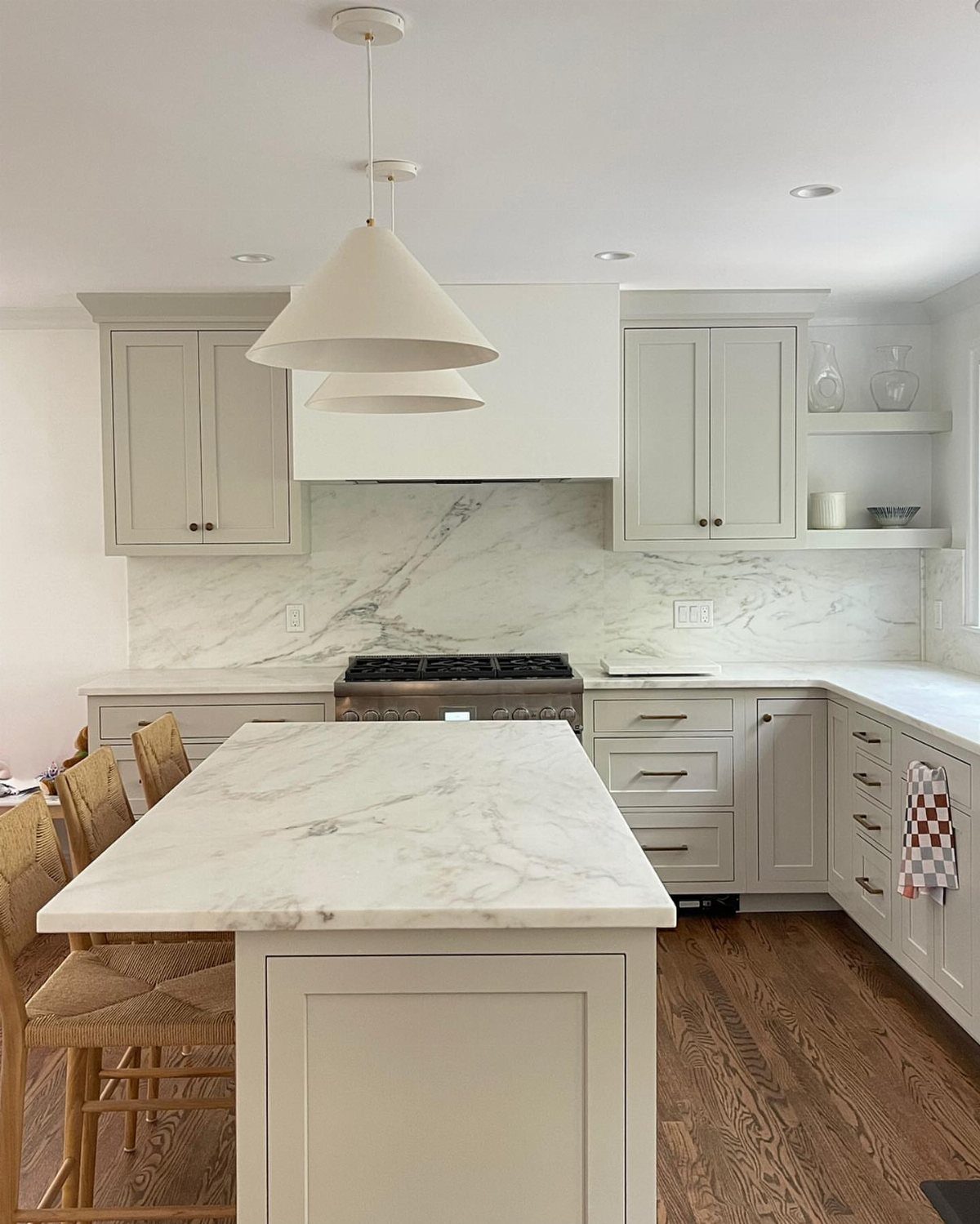 A bright kitchen with pale gray cabinets, marble countertops, and a marble backsplash with pendant lights.