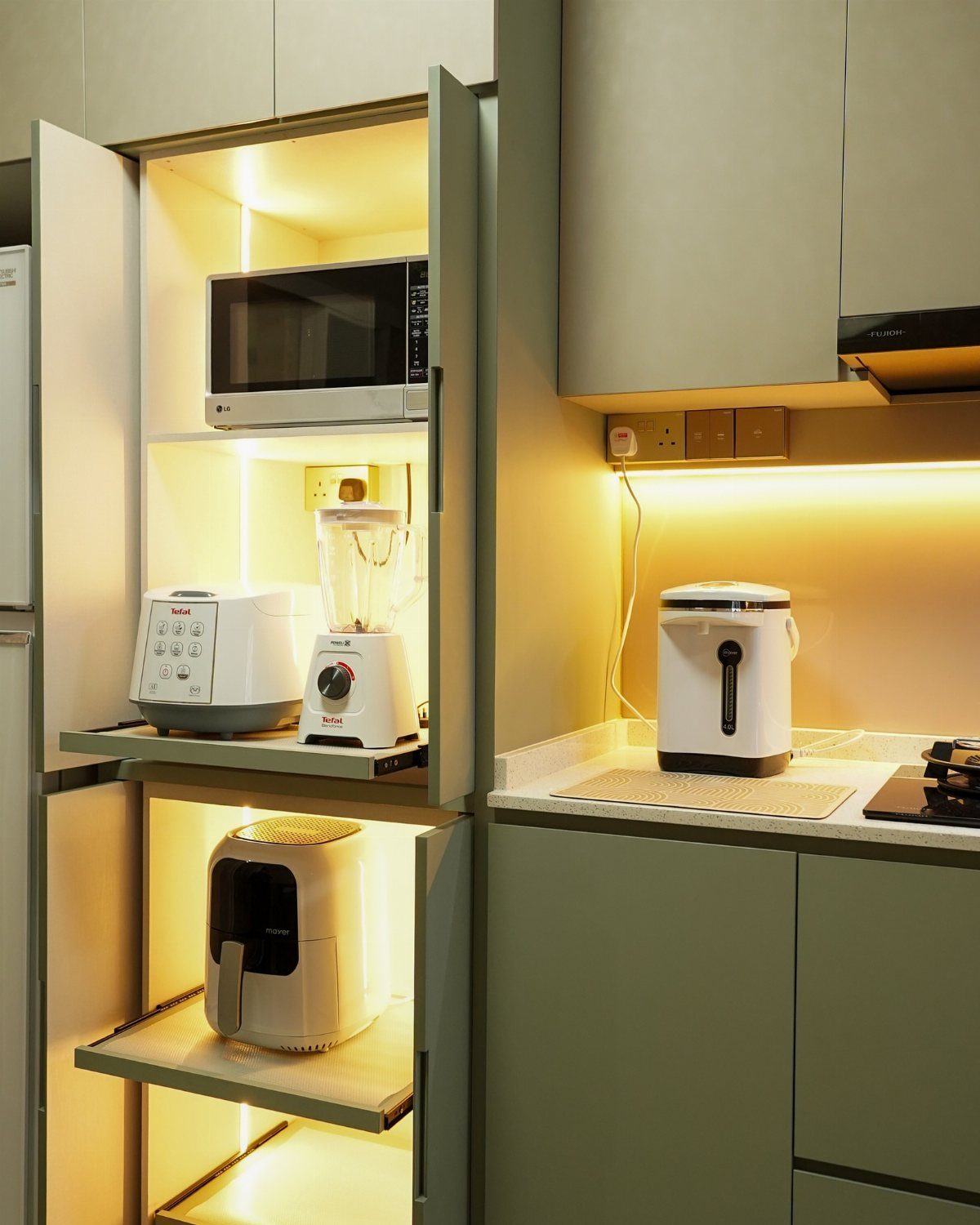 Small kitchen appliances organized inside a lit sage green cabinet and on a countertop.