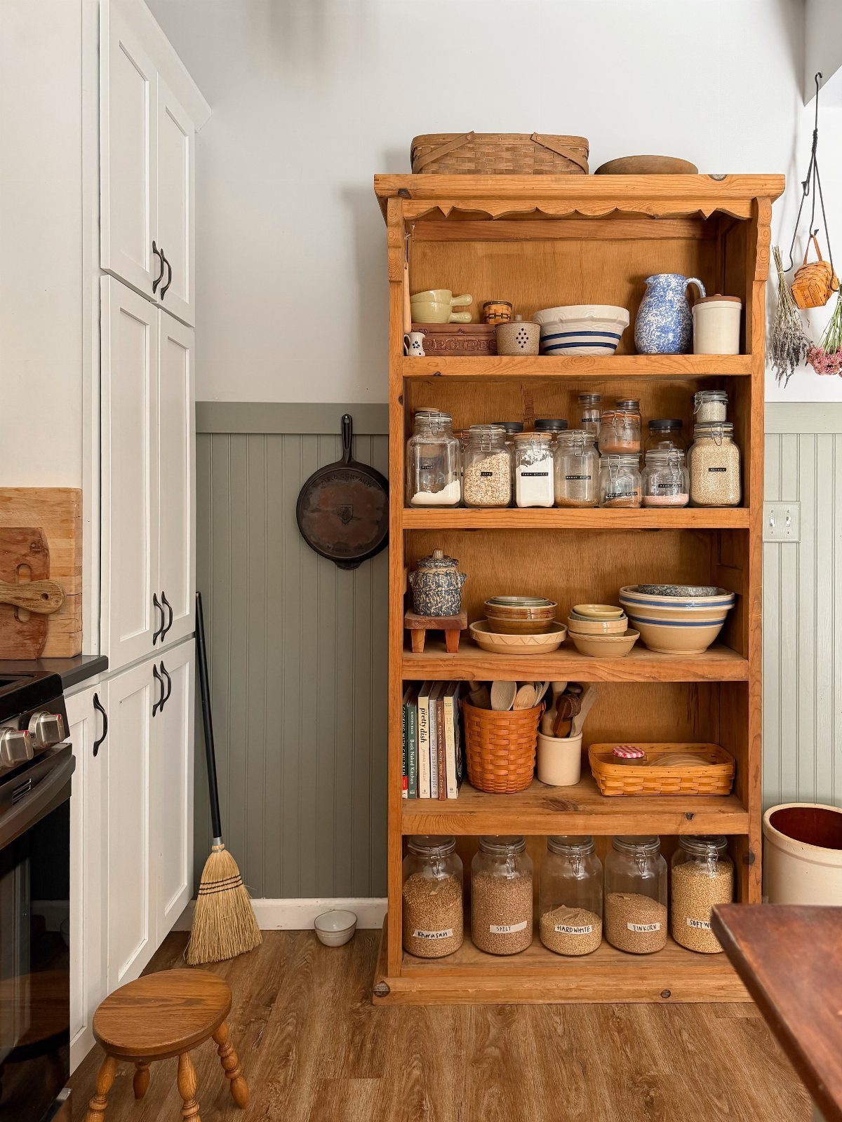 A wooden hutch with open shelves filled with jars, bowls, and kitchenware against a green wall.