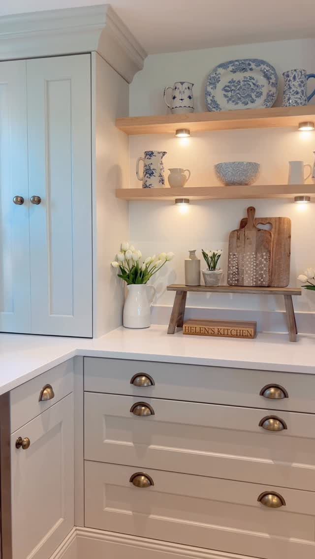 Light grey kitchen cabinets with brass hardware next to wooden floating shelves holding blue and white ceramics.