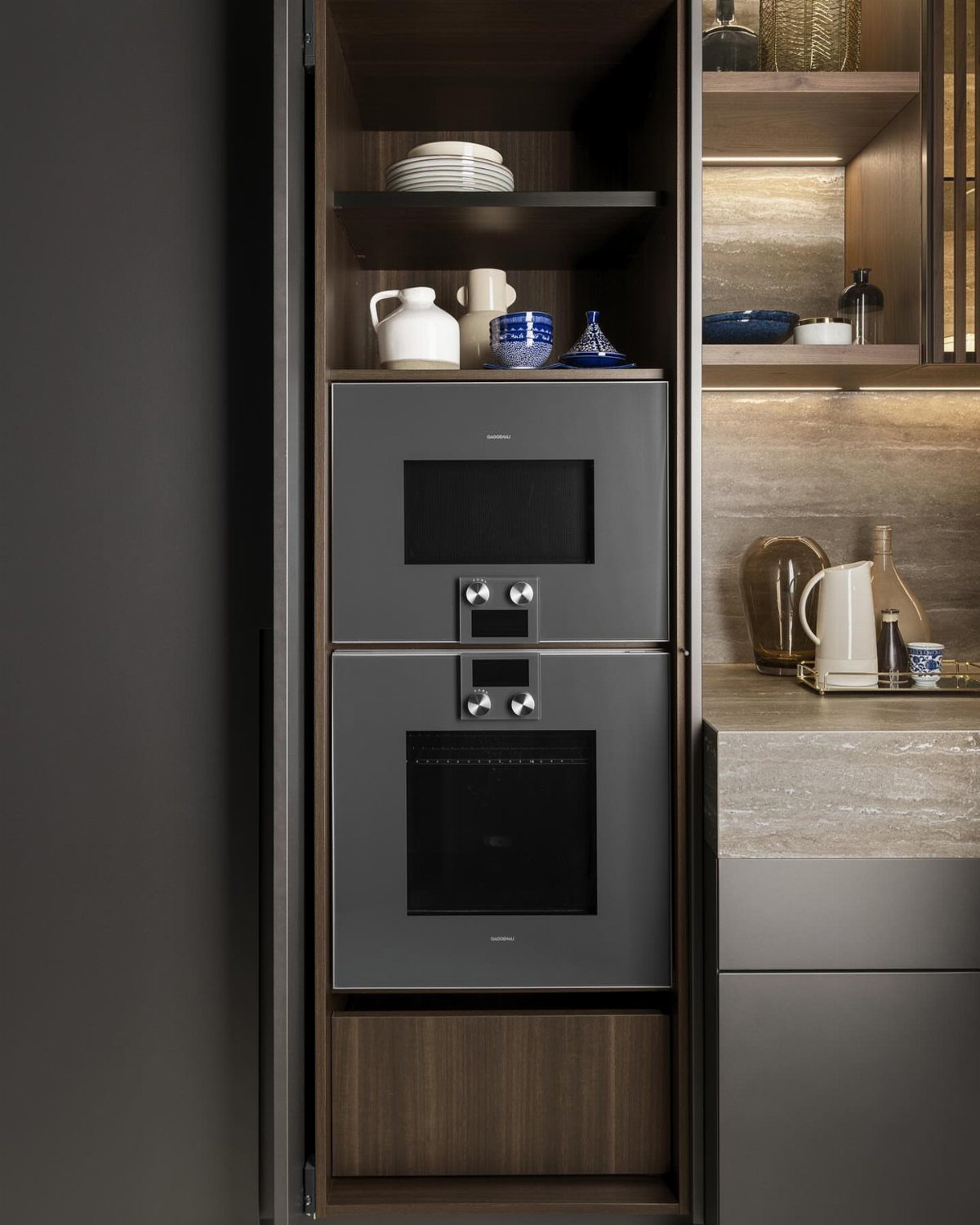 Stacked built-in ovens in a dark wood kitchen cabinet with open shelving above.