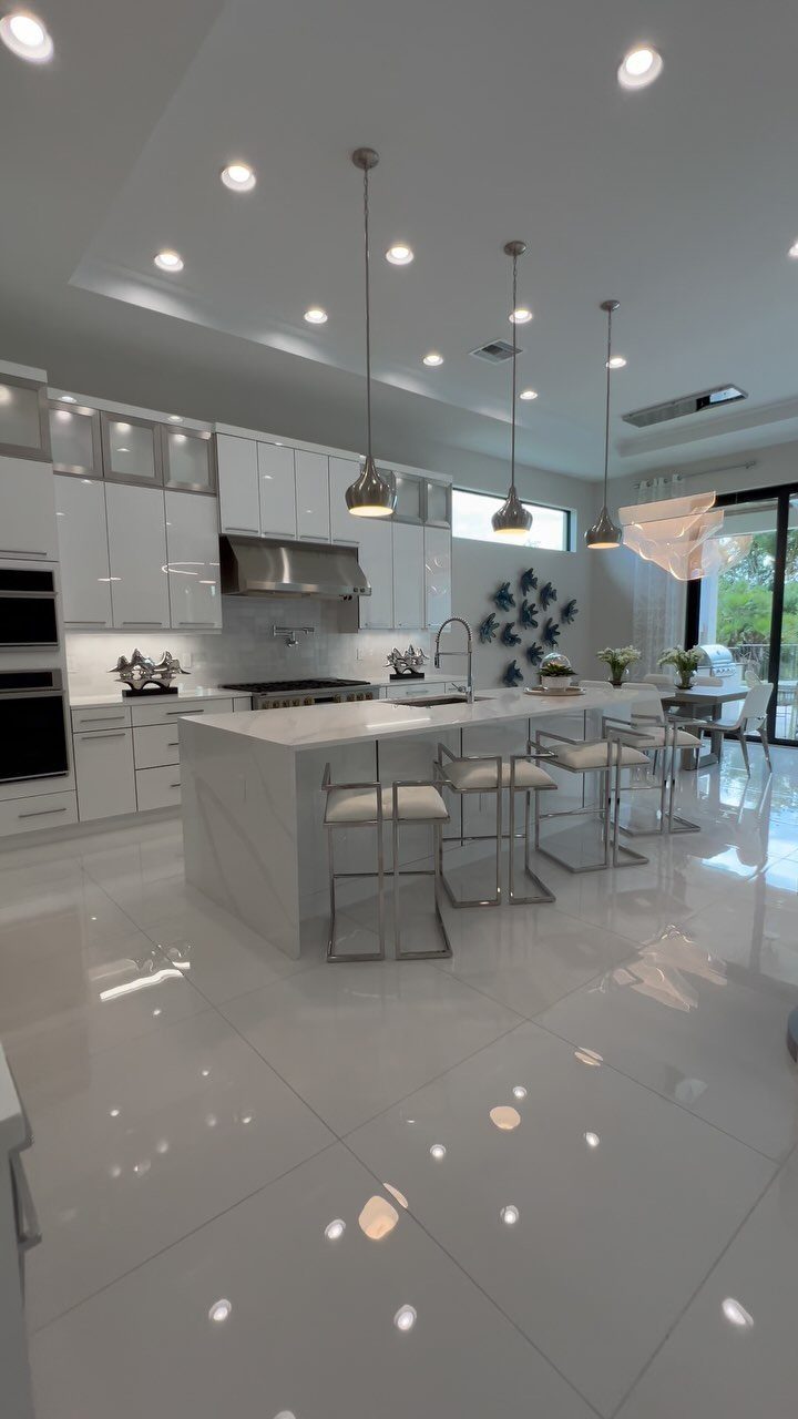 A modern white kitchen featuring a large marble island with four chrome barstools and pendant lighting.