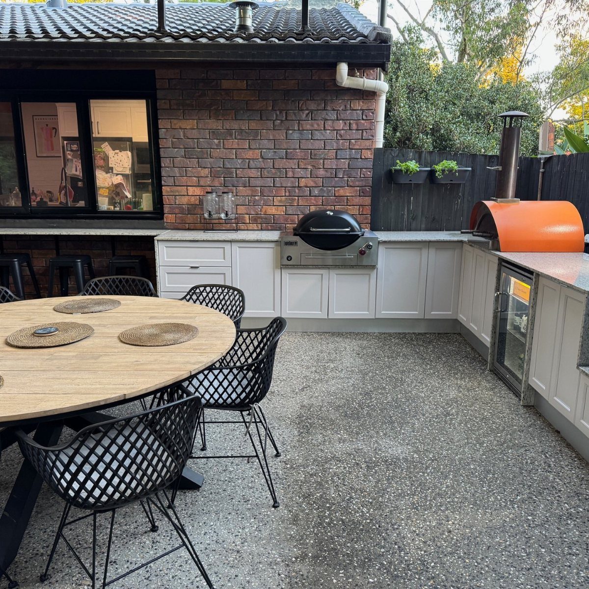 L-shaped outdoor kitchen with grey cabinets, a grill, and an orange pizza oven on a patio.