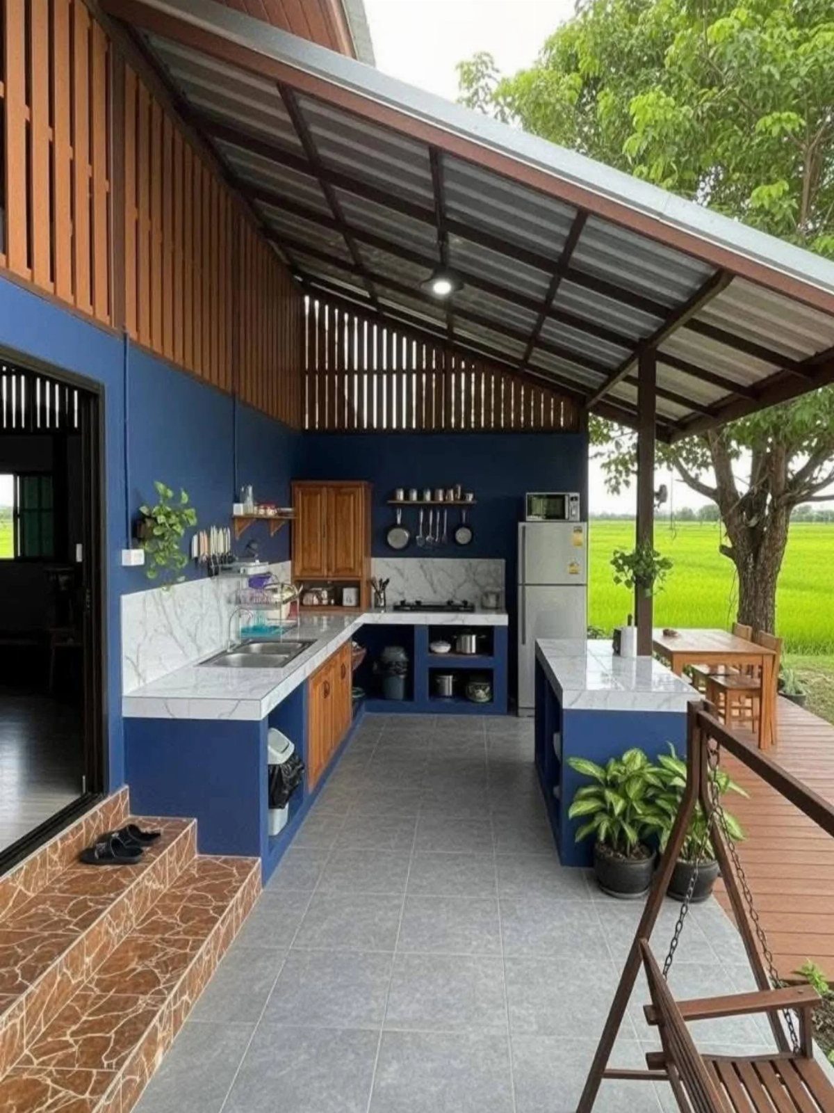 An L-shaped outdoor kitchen with blue cabinets and marble countertops under a covered patio area.