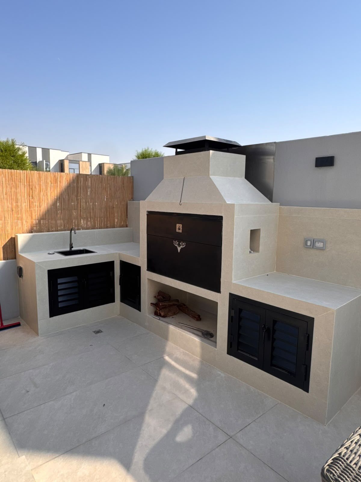 L-shaped outdoor kitchen with beige masonry, a black built-in grill, sink, and black cabinets.