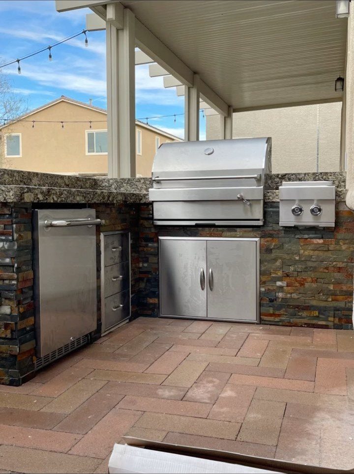 Stainless steel outdoor kitchen appliances built into a stone L-shaped counter under a patio.