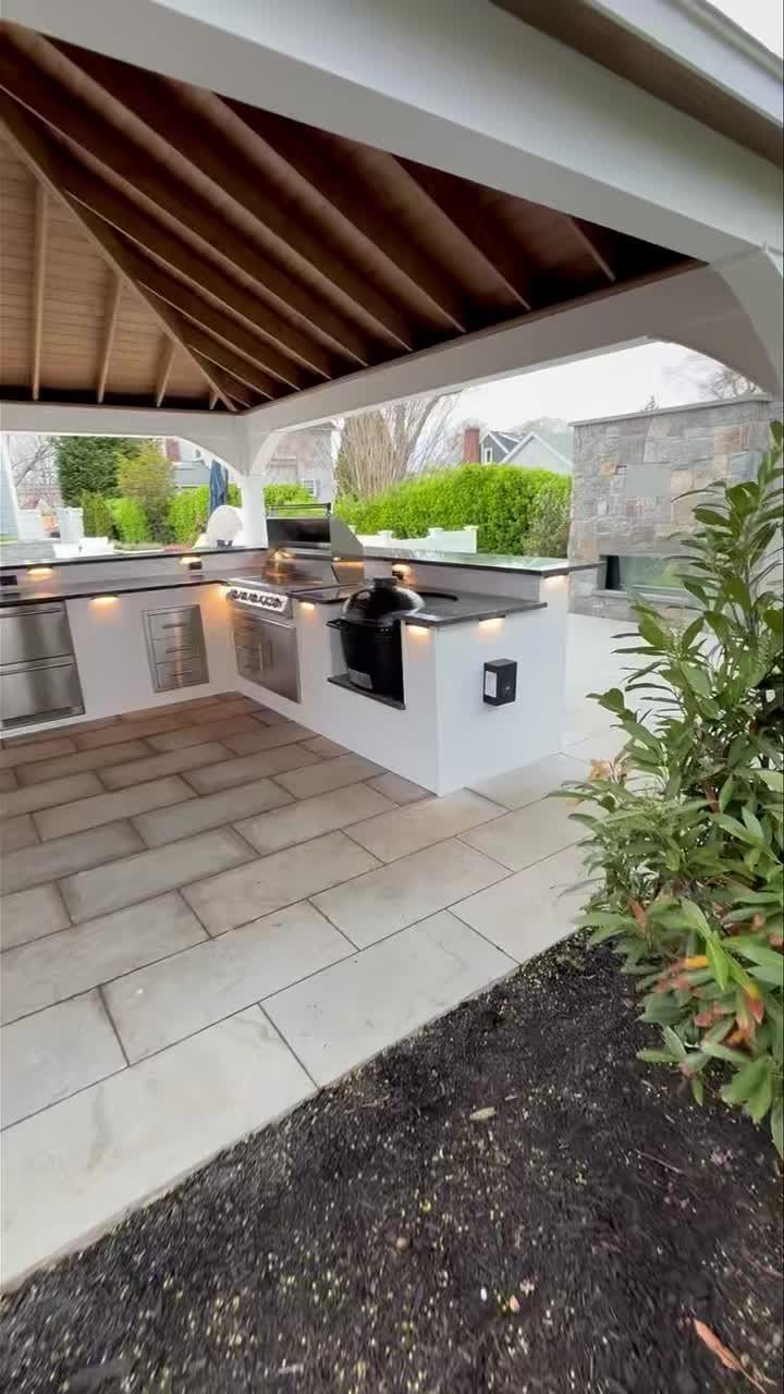 L-shaped outdoor kitchen with white cabinets and dark countertops under a wooden pavilion structure.