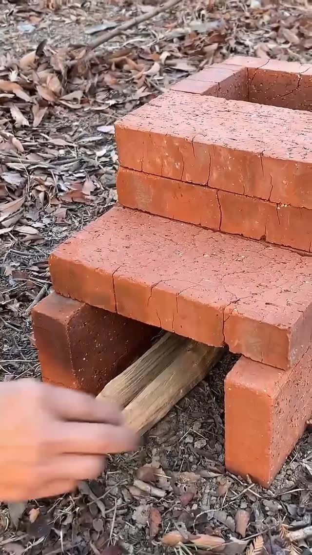 A hand placing wooden planks into a structure built from stacked red bricks outdoors.