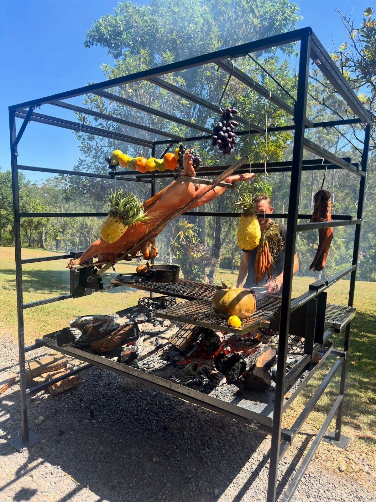 A large black metal frame structure outdoors cooking a whole pig and various fruits over fire.