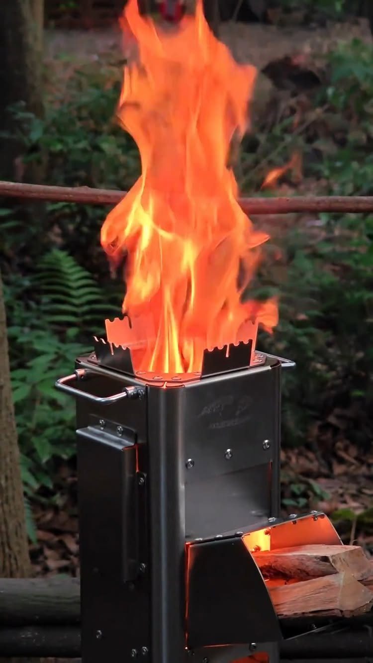 A metal rocket stove with bright orange flames rising from the top in a forest setting.