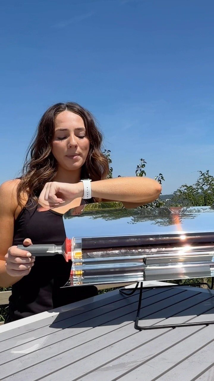 A woman checks her watch next to a reflective solar oven on an outdoor table.
