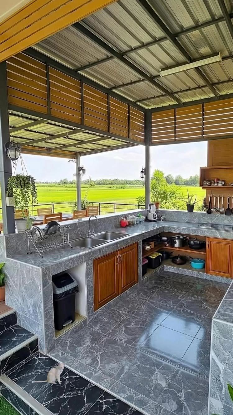 Outdoor kitchen with marble countertops, wooden cabinets, and a metal roof overlooking a green field.